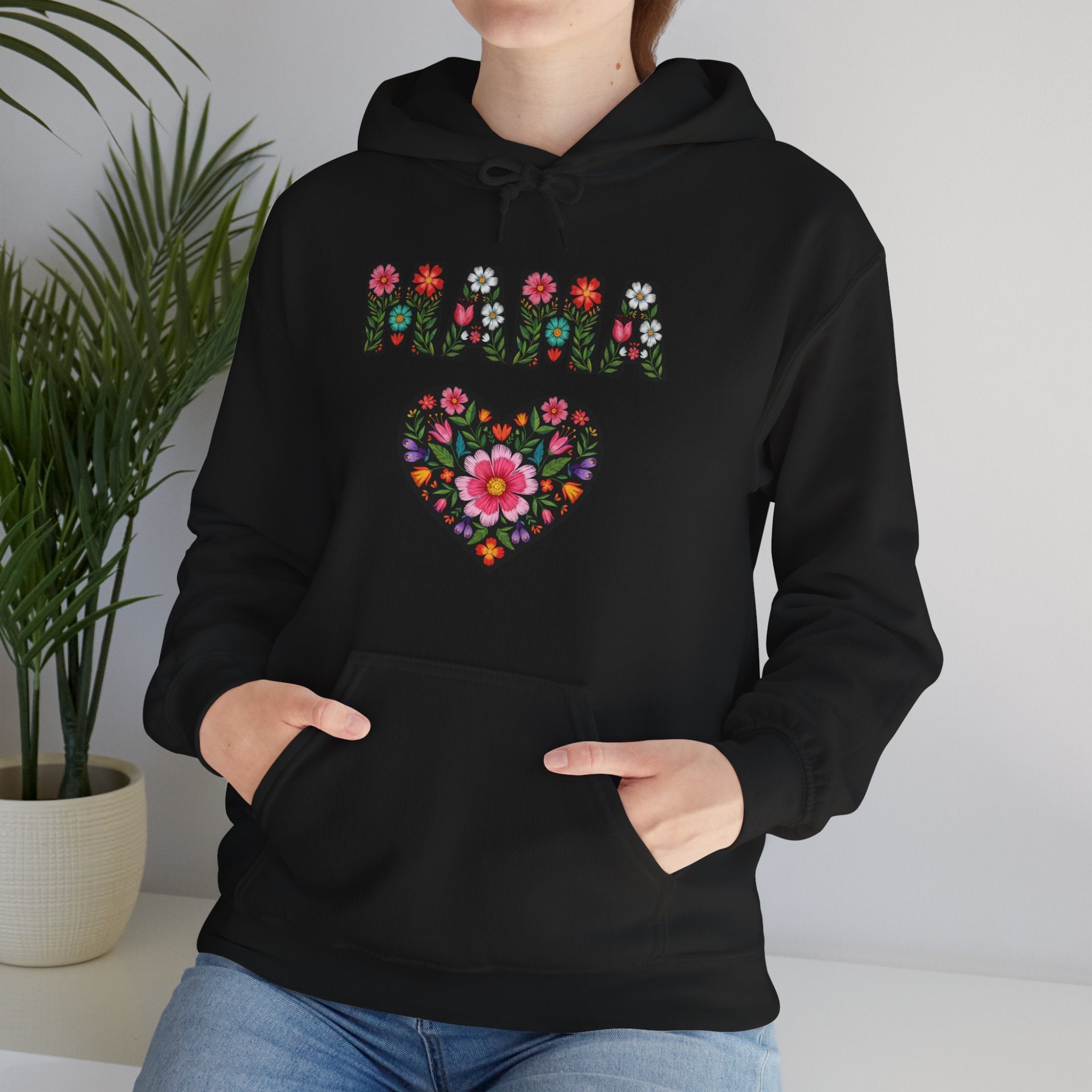 Person wearing a black hoodie with a colorful floral heart design, standing indoors next to a plant.