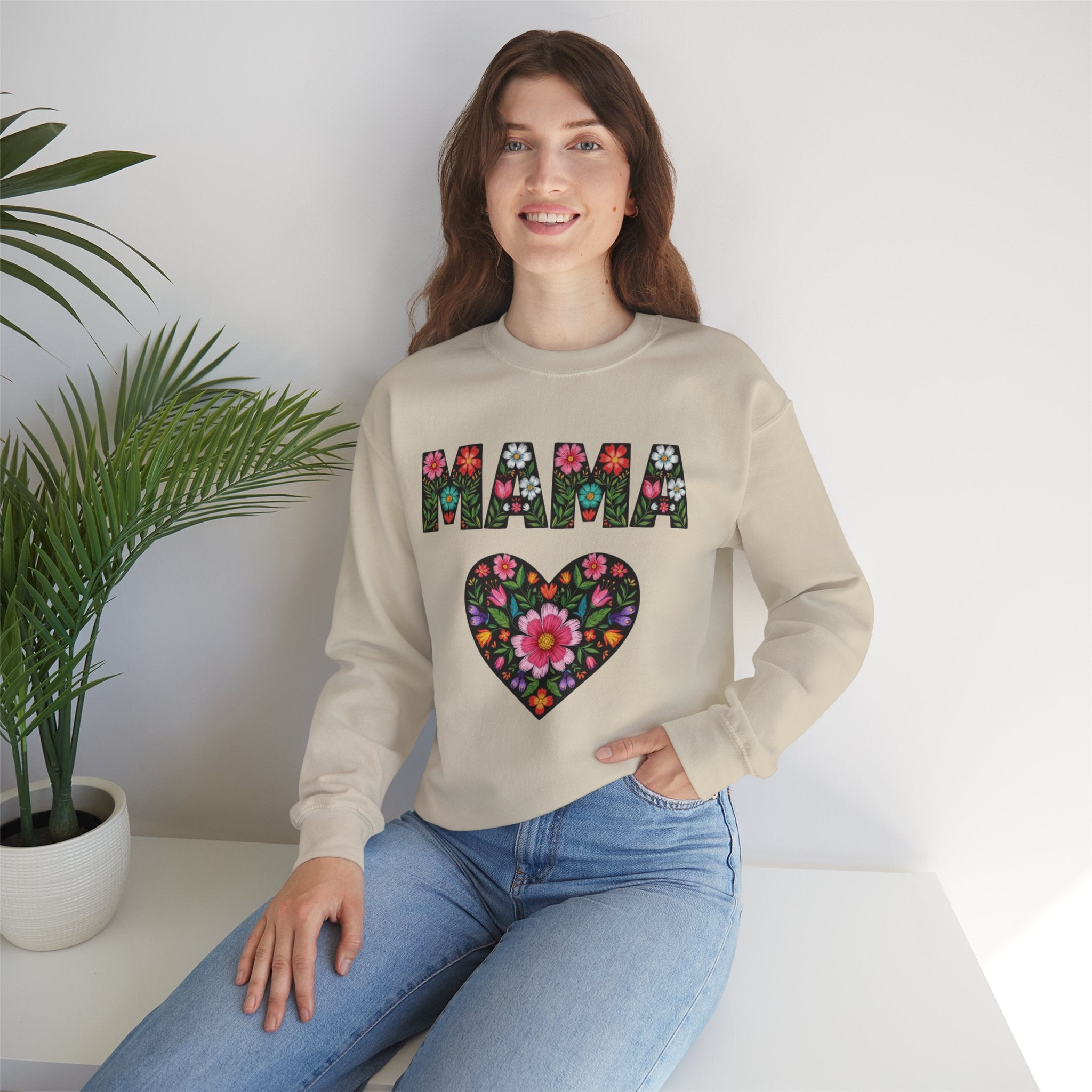 Woman wearing a beige sweatshirt with floral 'MAMA' design sitting on a white surface with a plant in the background.
