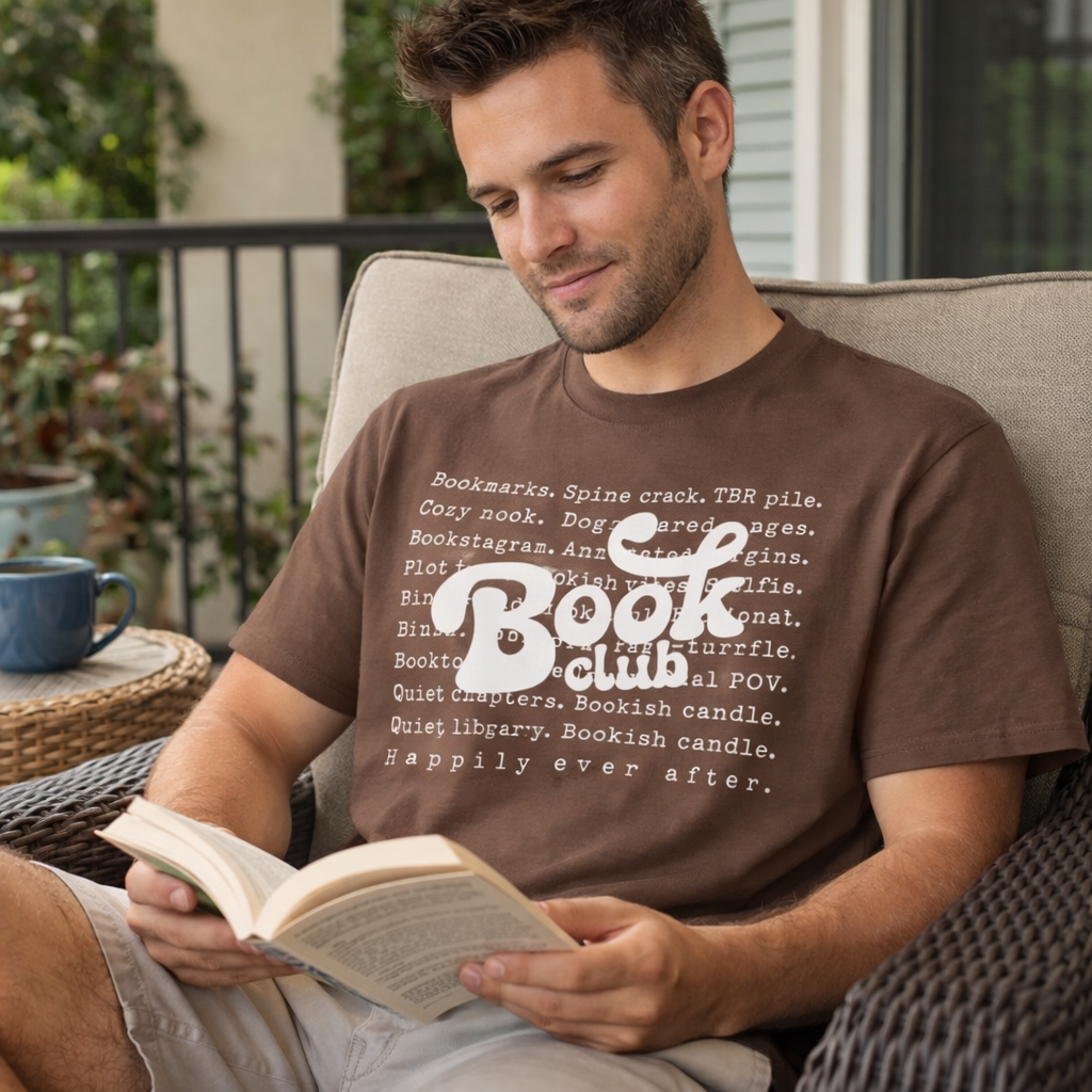 Man reading a book wearing a brown 'Book Club' t-shirt on a porch.